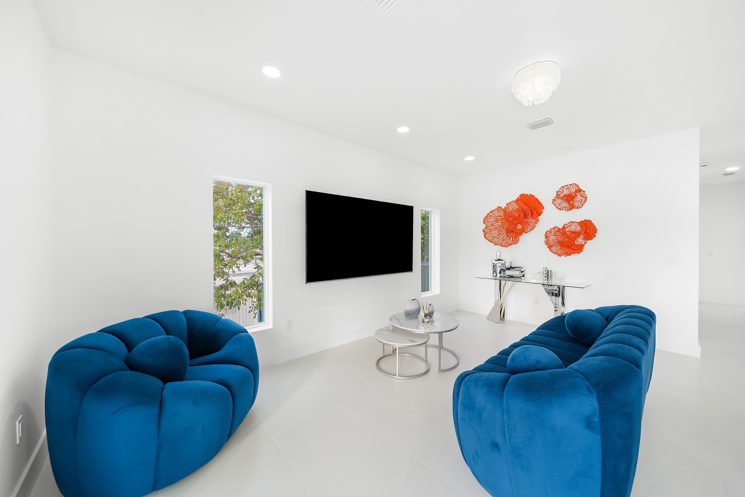 Een moderne woonkamer met een flatscreen-tv, twee blauwe banken, een klein rond tafeltje en decoratieve bloemenkunst op een oranje muur.