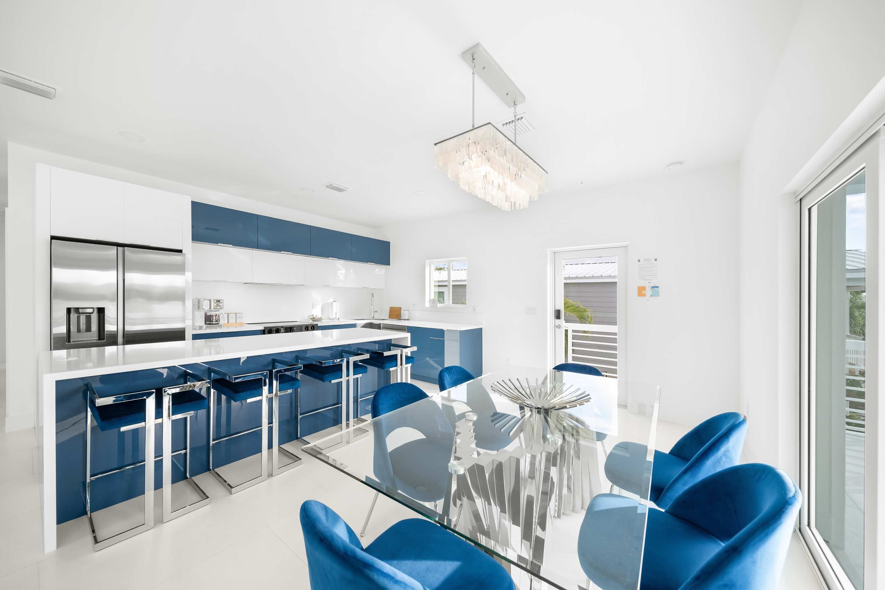 A modern kitchen with a central island, bar stools, and a dining area with blue chairs and a glass table.