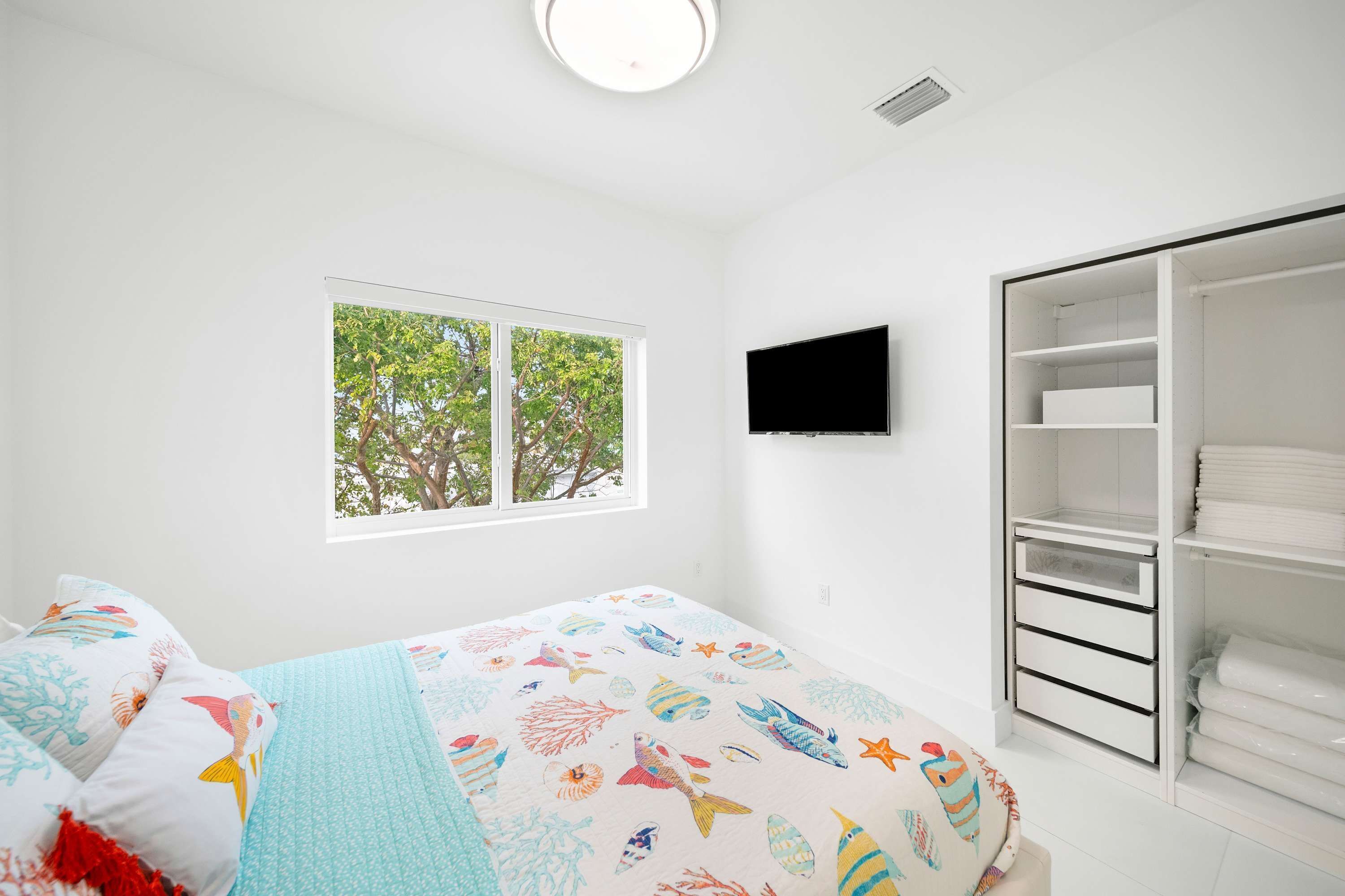 A bedroom with a bed, a window with a view of trees, a TV mounted on the wall, and a white wardrobe.