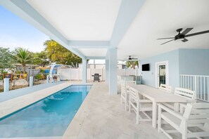 Un espace piscine extérieur abrité, avec une table et des chaises blanches, un ventilateur de plafond et une vue sur le jardin arrière.