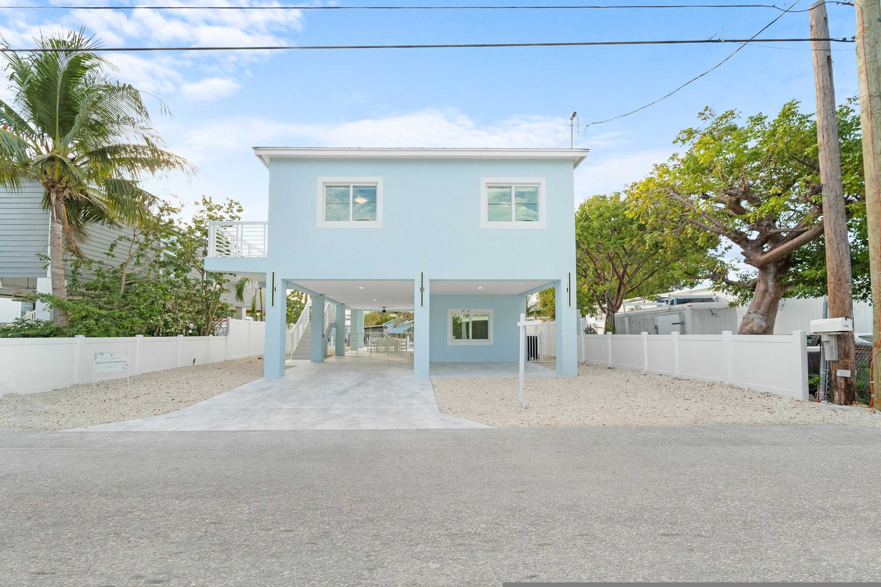 Une maison bleu clair à deux étages, dotée d’un porche abrité, entourée d’une clôture blanche, de palmiers et surplombant un ciel dégagé.