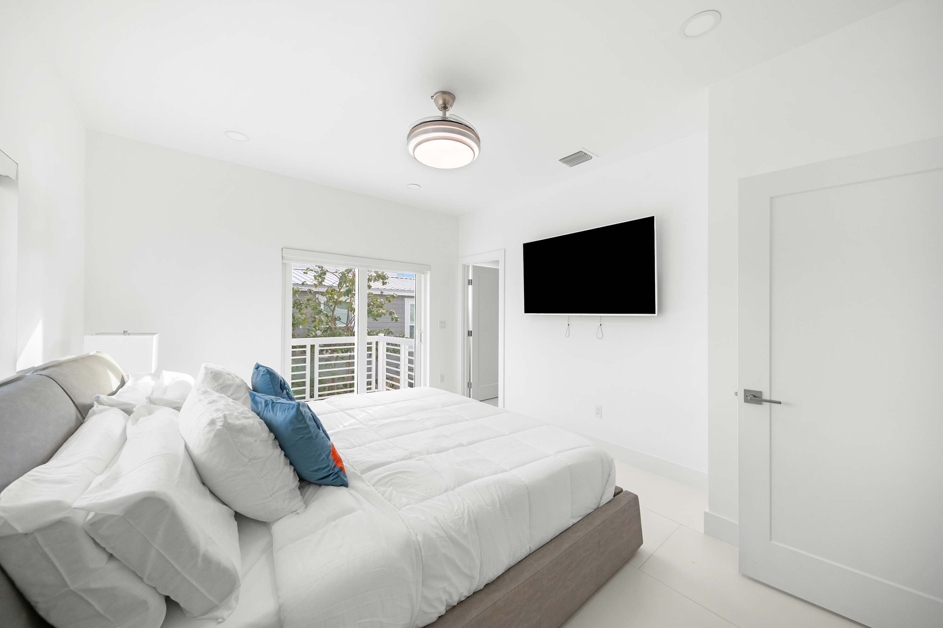 A bedroom with a bed, white walls, a ceiling fan, a TV mounted on the wall, and a door.
