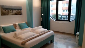A neatly made bed with white linens and a brown throw, a painting on the wall, and a view of buildings through a window with green curtains. - Room Kabine 1 (Koksijde)