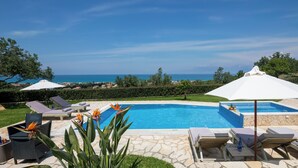 Une piscine entourée de verdure, avec des chaises longues et des parasols, et offrant une vue sur la mer.