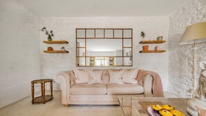 A living room with a beige sofa, a wooden coffee table, and a mirror above the sofa. There are wooden shelves with plants and decorative items on the walls.