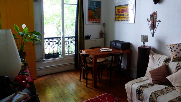 A room with a wooden dining table, chairs, a sofa, a radiator, a window with curtains, a plant, and a poster on the wall. - Charming apartment in Belleville (Paris)