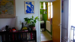 A room with a bookshelf, a map, a framed abstract painting, and a view into a kitchen. - Charming apartment in Belleville (Paris)