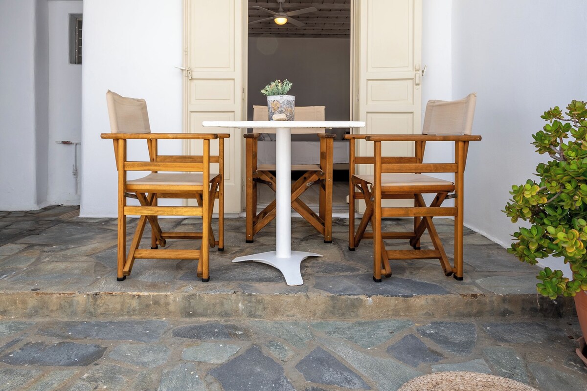 A patio with a white table and four wooden chairs, a stone floor, and a white ceiling with wooden beams.