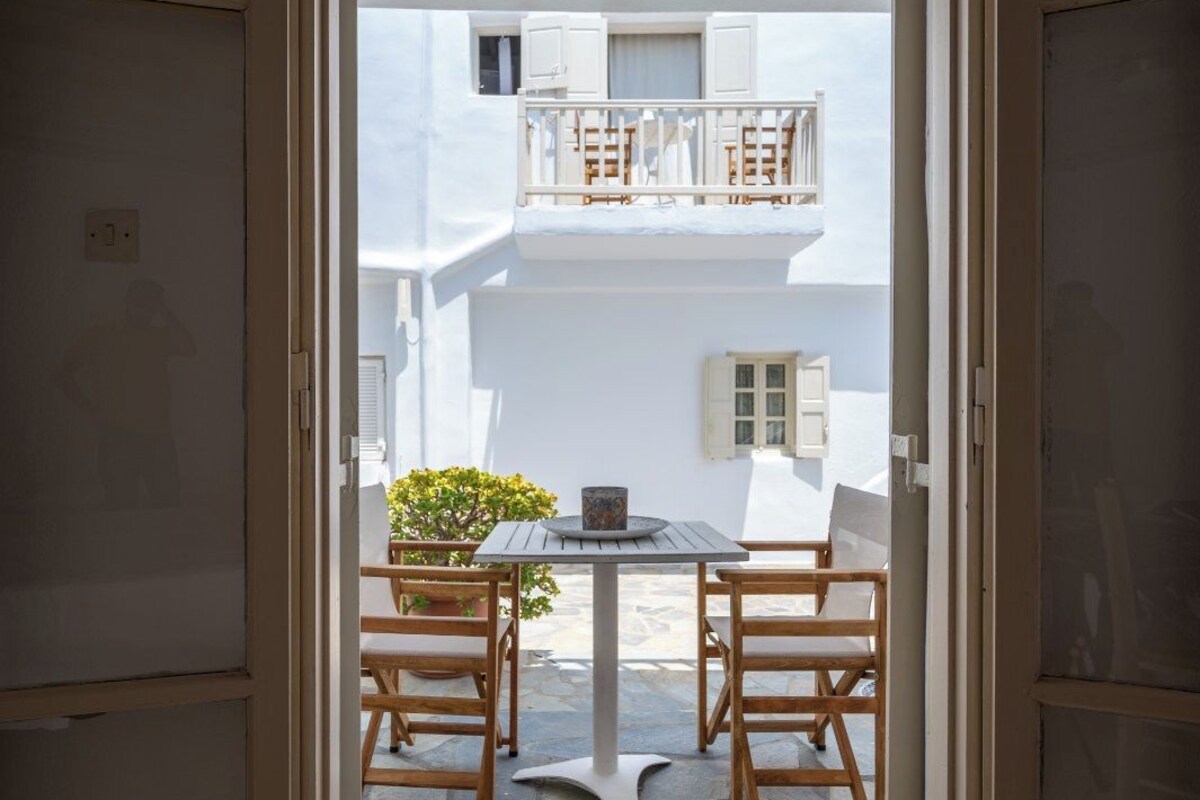 An open doorway leading to a stone-floored patio with a table and chairs, a potted plant, and a white balcony with shutters.