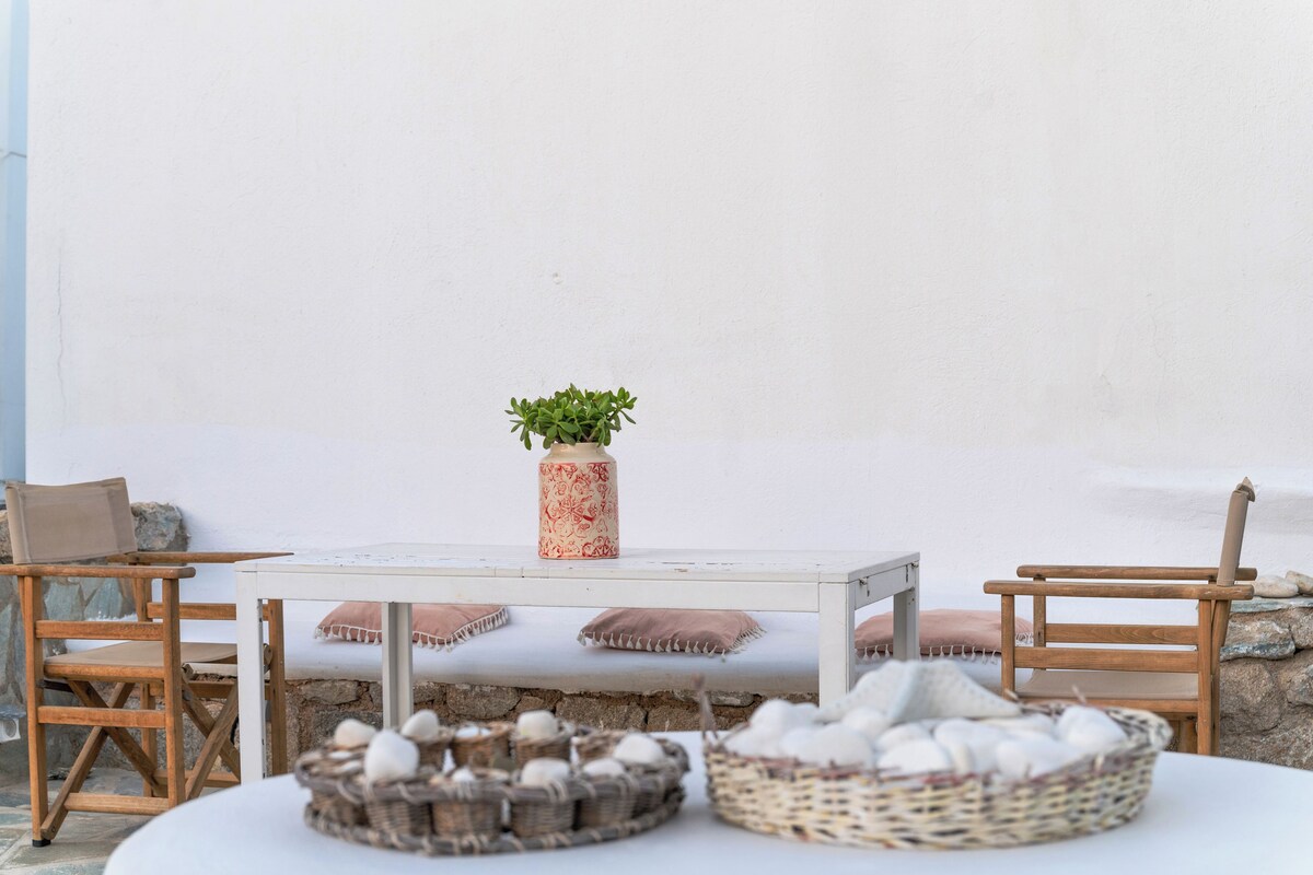 Una mesa blanca con un florero de plantas verdes, dos sillas de madera y una cesta de huevos sobre un suelo de piedra.