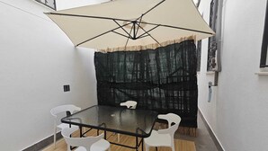 Un petit espace extérieur aménagé pour les repas, à l’abri, avec une table et quatre chaises, un grand parasol et un fond noir texturé.