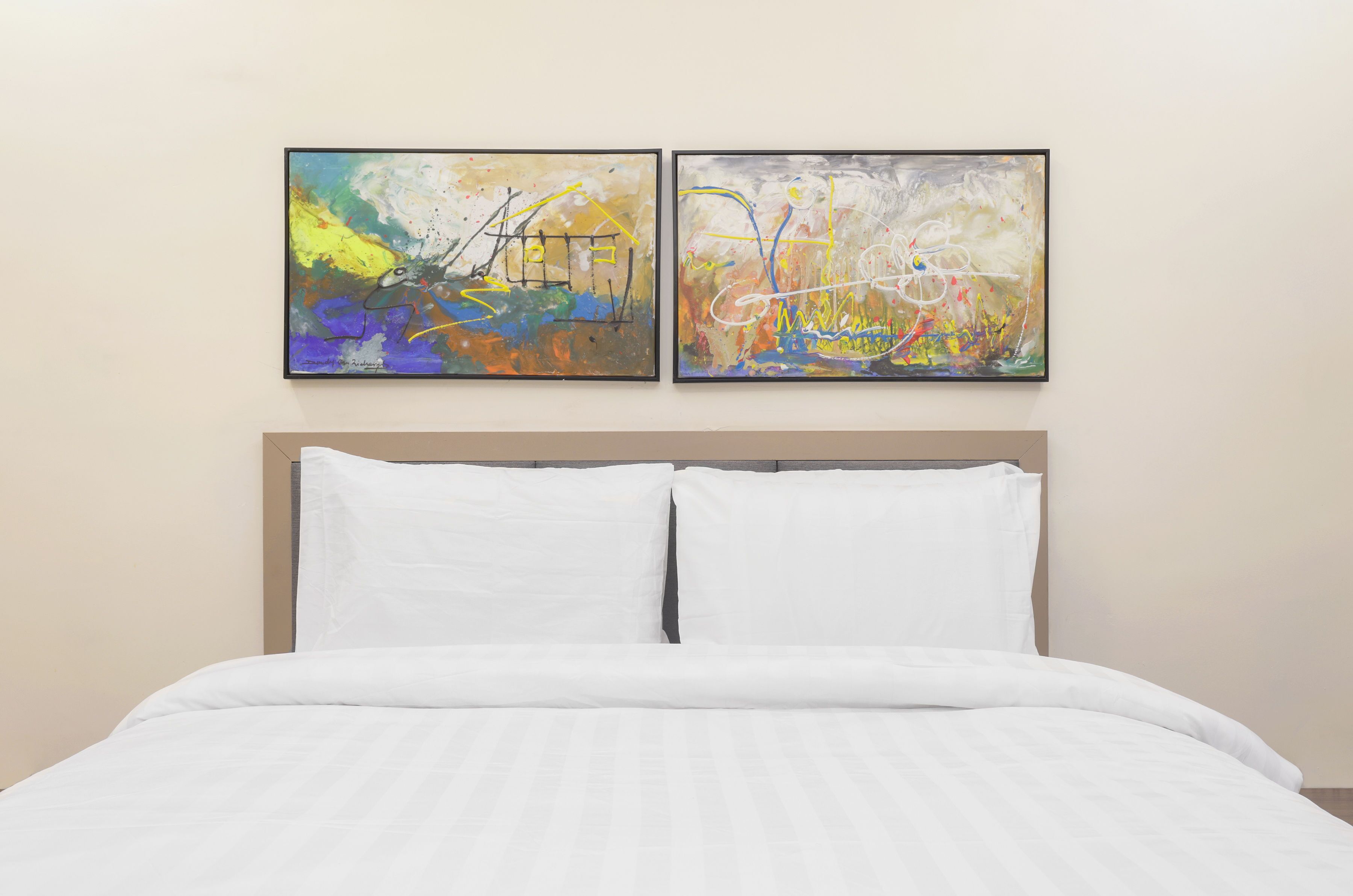 Una cama con ropa blanca y dos cuadros abstractos sobre ella.