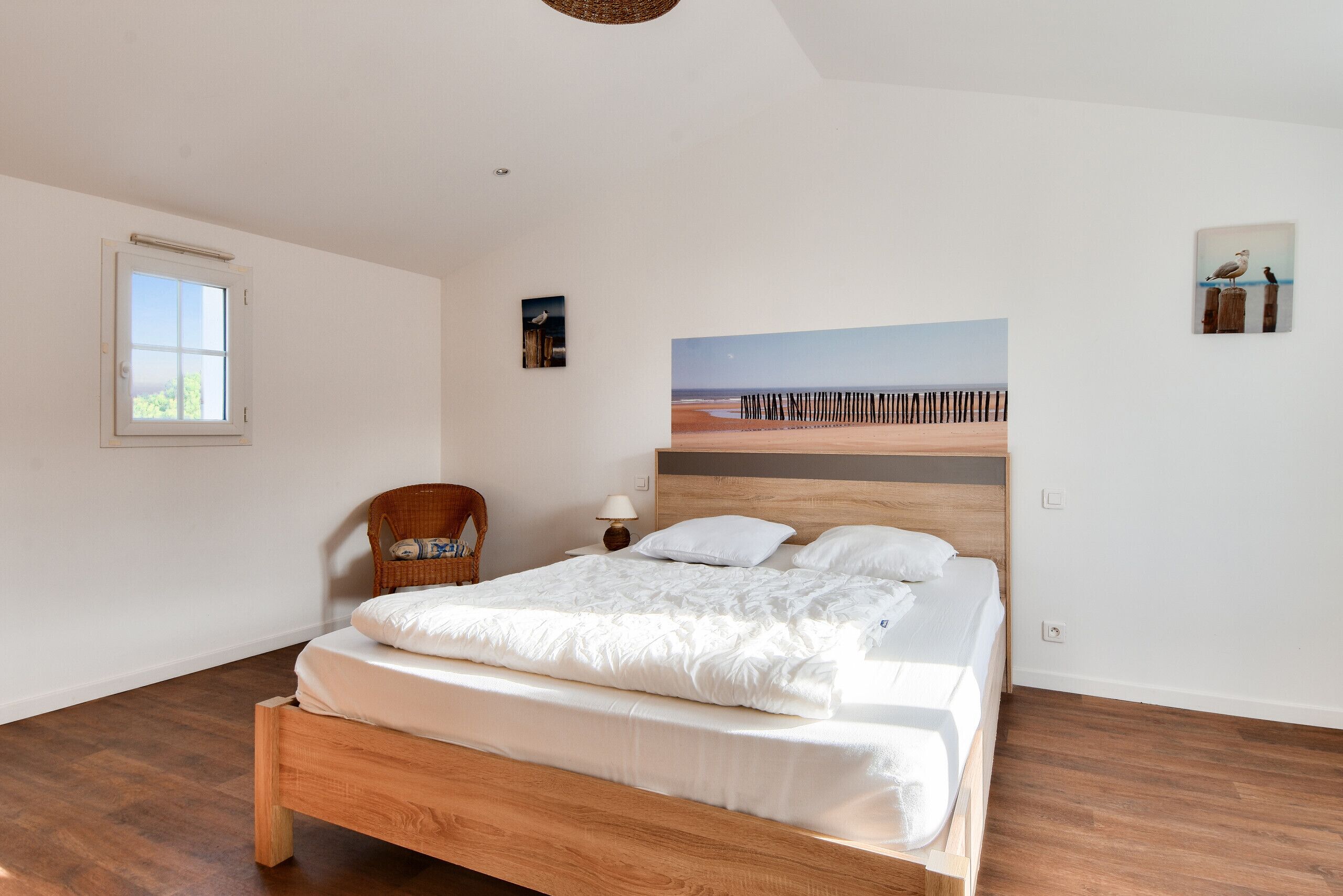 A bedroom with a wooden bed, a wicker chair, a painting of a beach, and a window with blinds.