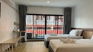 A hotel room with two beds, a desk, a chair, and a large window with a view of a red building. - M3 Bangkok Bake & Bed (Bangkok)