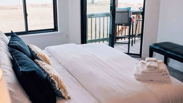 A bedroom with a bed, pillows, a stack of towels, and a view of the beach through a large window. - Artist Windmill Pt Condo Sunset Pool Relaxation (White Stone)