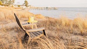 A beach chair with a yellow container on a sandy beach with tall grass and a pier in the background. - Artist Windmill Pt Condo Sunset Pool Relaxation (White Stone)