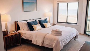 A neatly made bed with white and blue bedding, a bedside lamp, and a view of the beach through a large window. - Artist Windmill Pt Condo Sunset Pool Relaxation (White Stone)