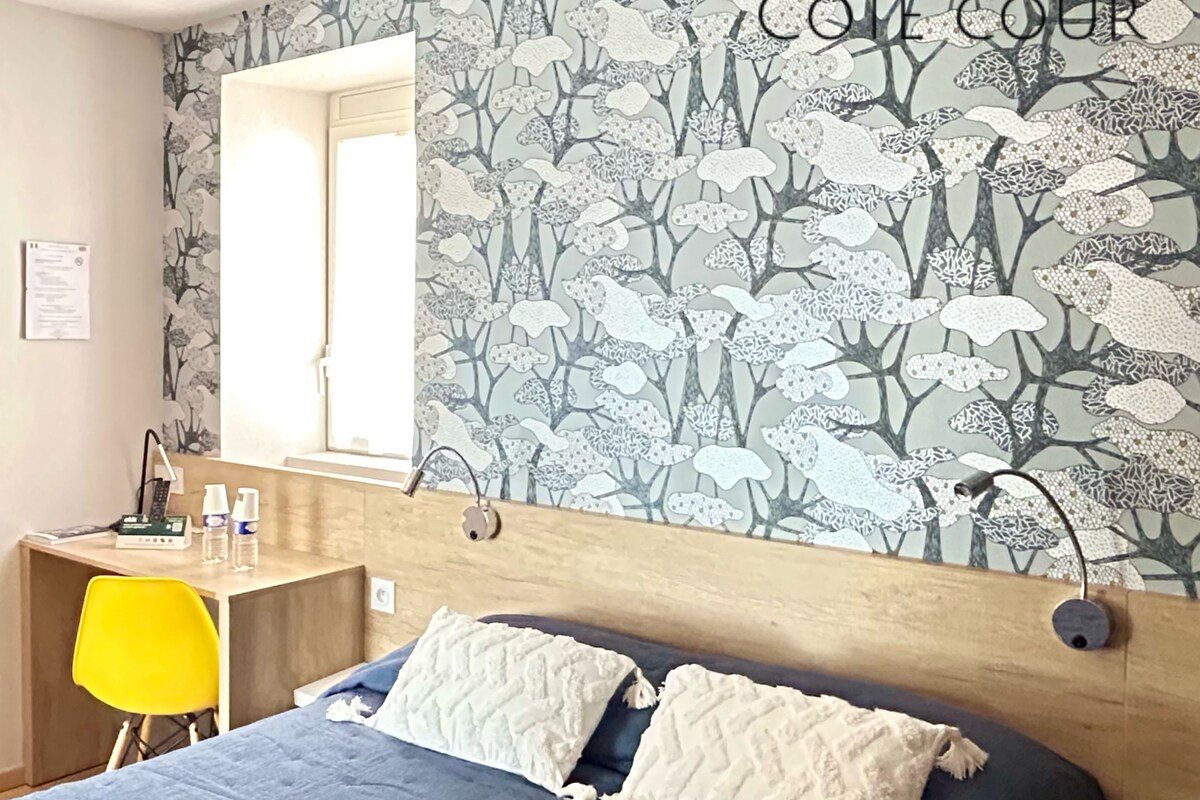 A bedroom with a bed, a desk, and a chair. There is a window and a wall with a floral pattern.