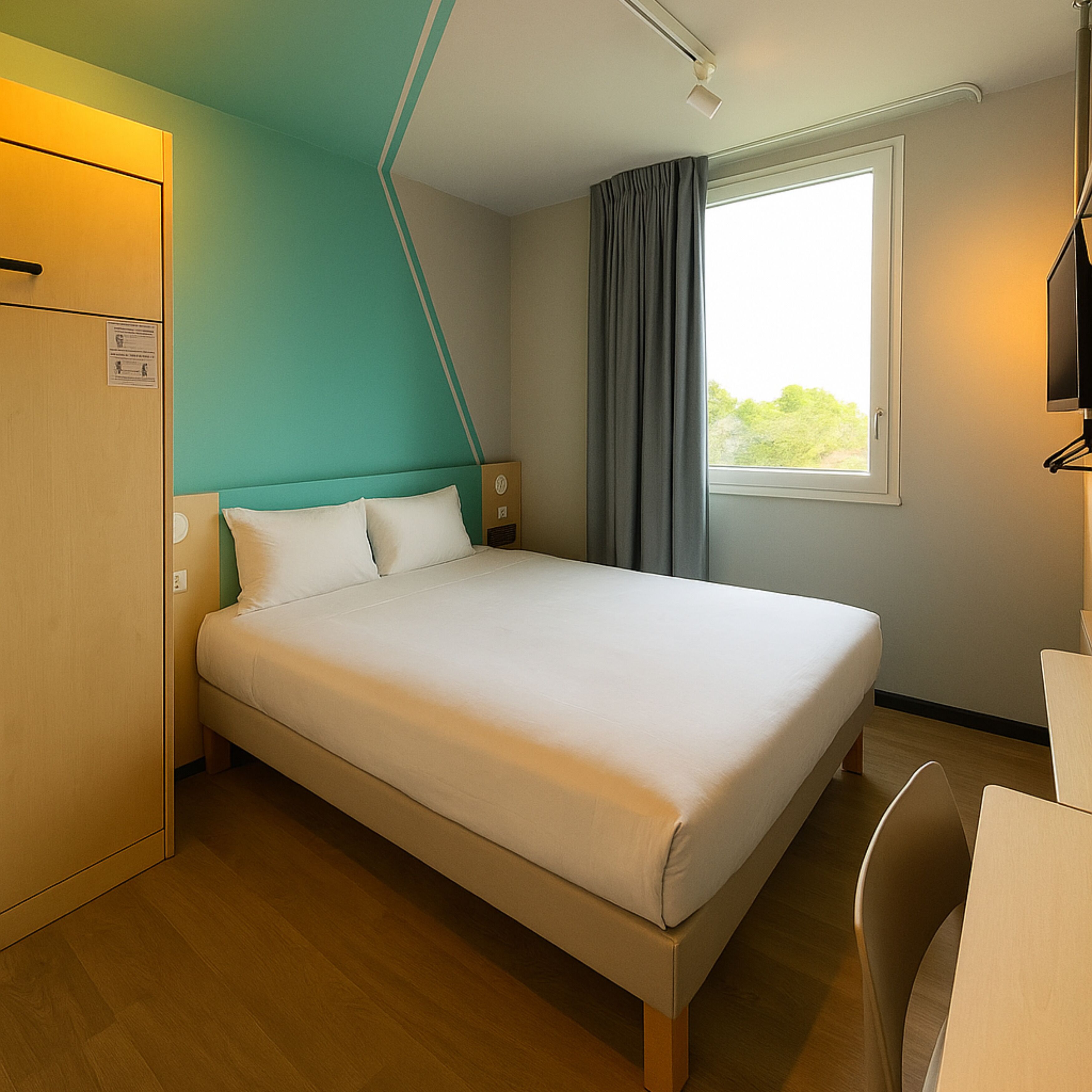 A hotel room with a bed, a desk, a chair, a wardrobe, a window with curtains, and a television.