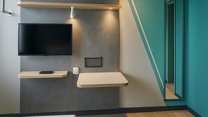 A modern room with a flat-screen TV mounted on a grey wall, wooden shelves, and a mirror reflecting the room. - Ibis Budget Auxerre Nord (Ouverture Printemps 2025) (Auxerre)