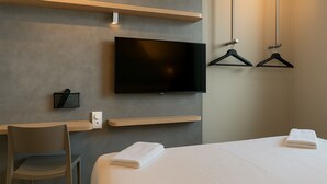A modern hotel room with a flat-screen TV mounted on the wall, a desk with a chair, and a bed with a neatly folded white towel. - Ibis Budget Auxerre Nord (Ouverture Printemps 2025) (Auxerre)