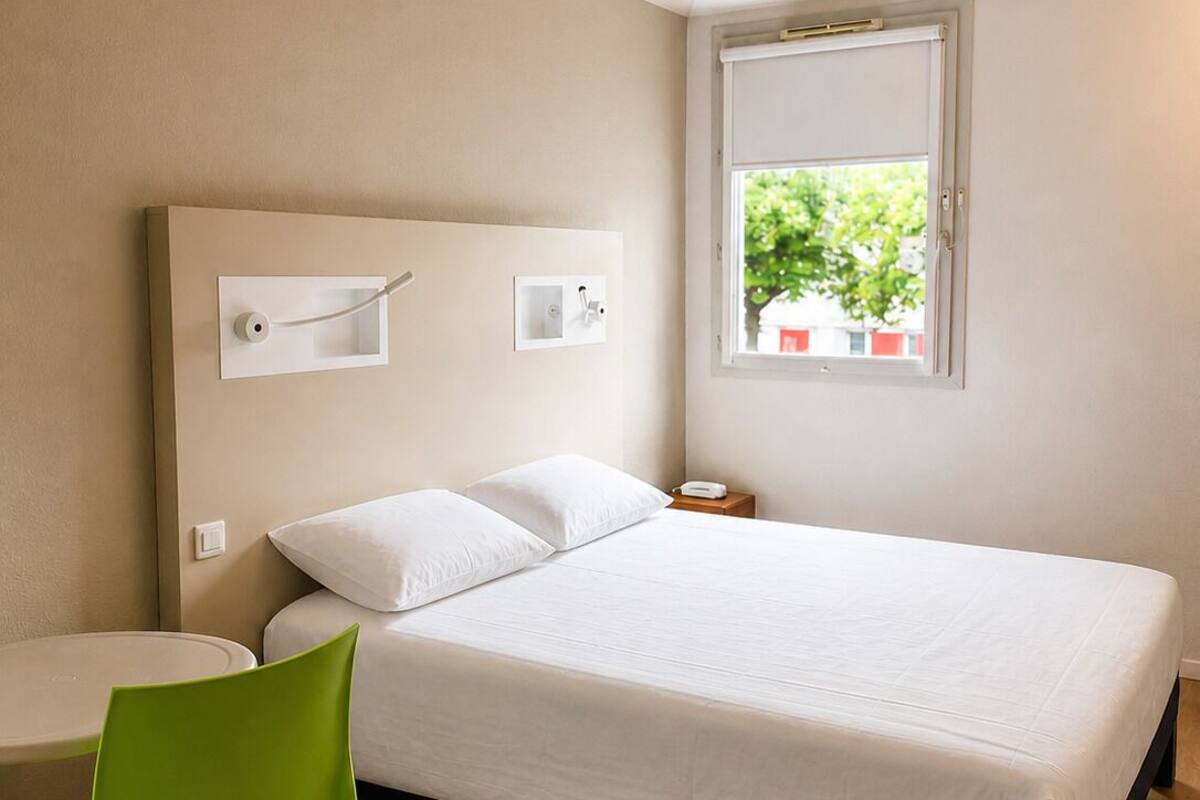 Een slaapkamer met een bed, een stoel, een nachtkastje, een raam en een wandlamp.