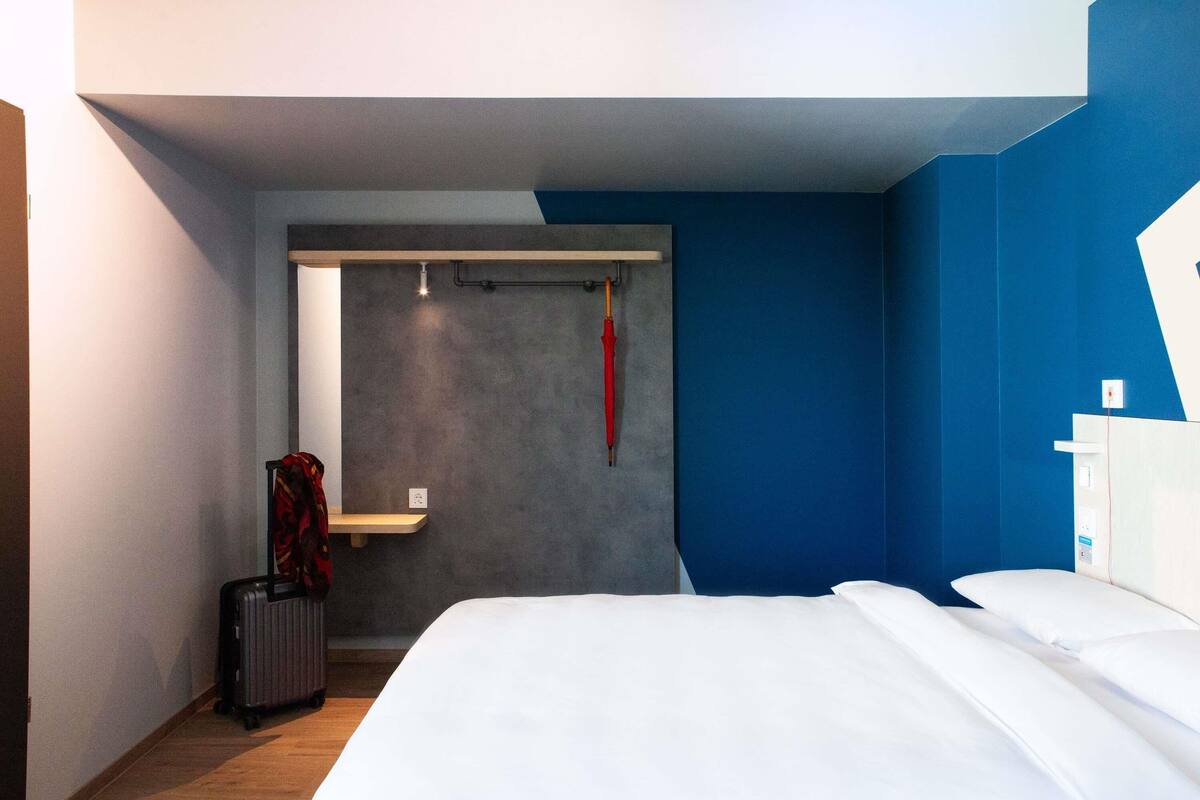 A hotel room with a bed, a suitcase, a coat rack, and a wall-mounted light fixture.
