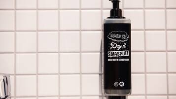 Une bouteille noire avec un distributeur à pompe, portant l'étiquette « LAVEZ-LE. SÉCHEZ-LE. ÉCRaseZ-LE ! GEL LAVANT POUR CHEVEUX, CORPS ET MAINS », est placée devant un mur carrelé.