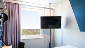 Habitación de hotel con cama, escritorio, silla, televisor y ventana con vista a un pueblo.