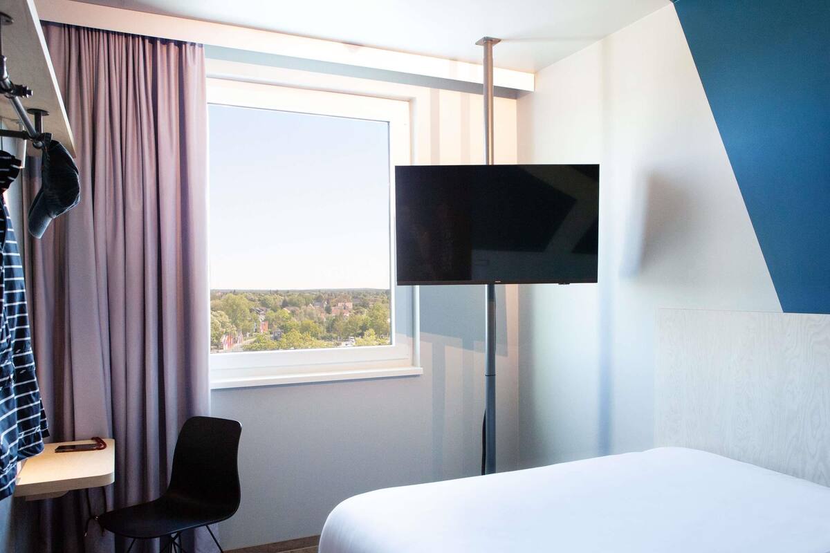 A hotel room with a bed, a desk, a chair, a TV, and a window with a view of a town.