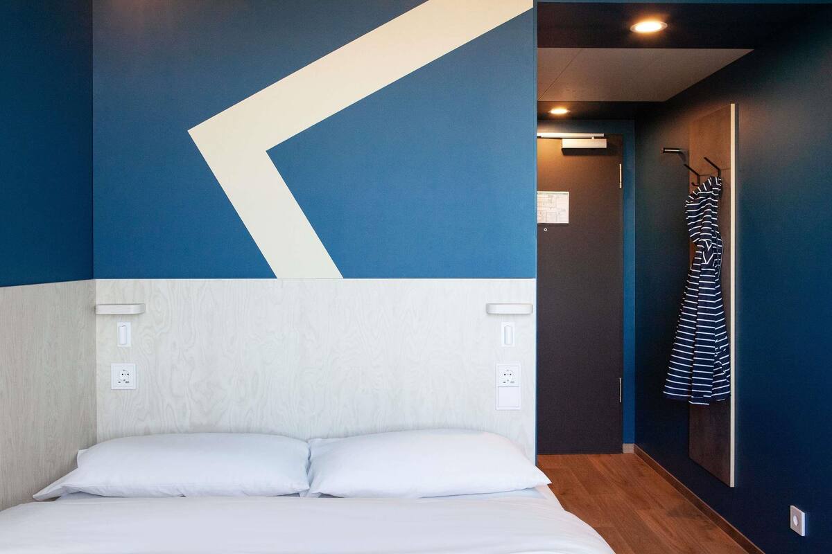 A hotel room with a bed, a wall with a geometric design, a coat rack, and a door leading to another room.