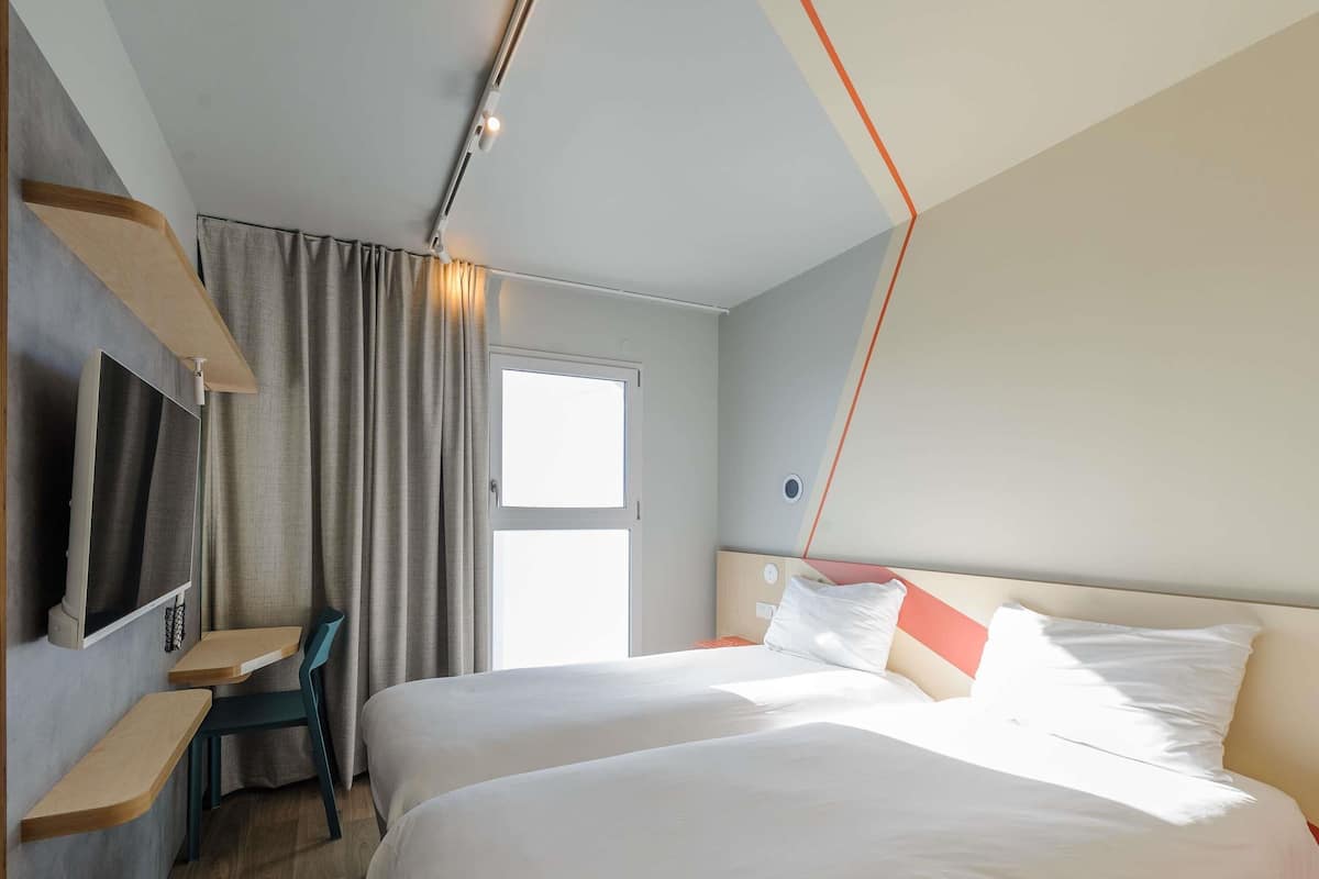 A hotel room with two beds, a desk, a TV, and a window.