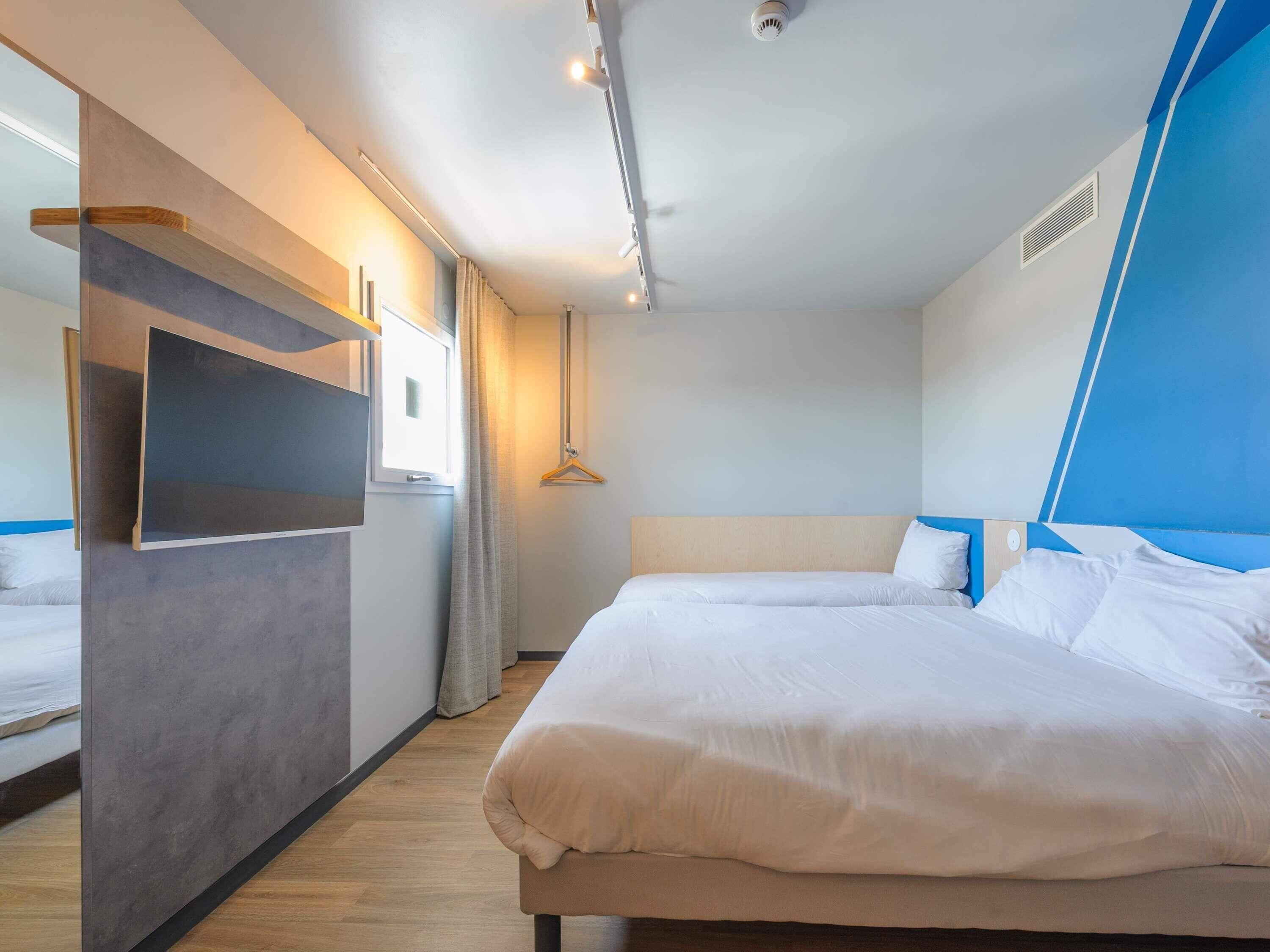 A modern hotel room with a large bed, a built-in TV, and a window with curtains.