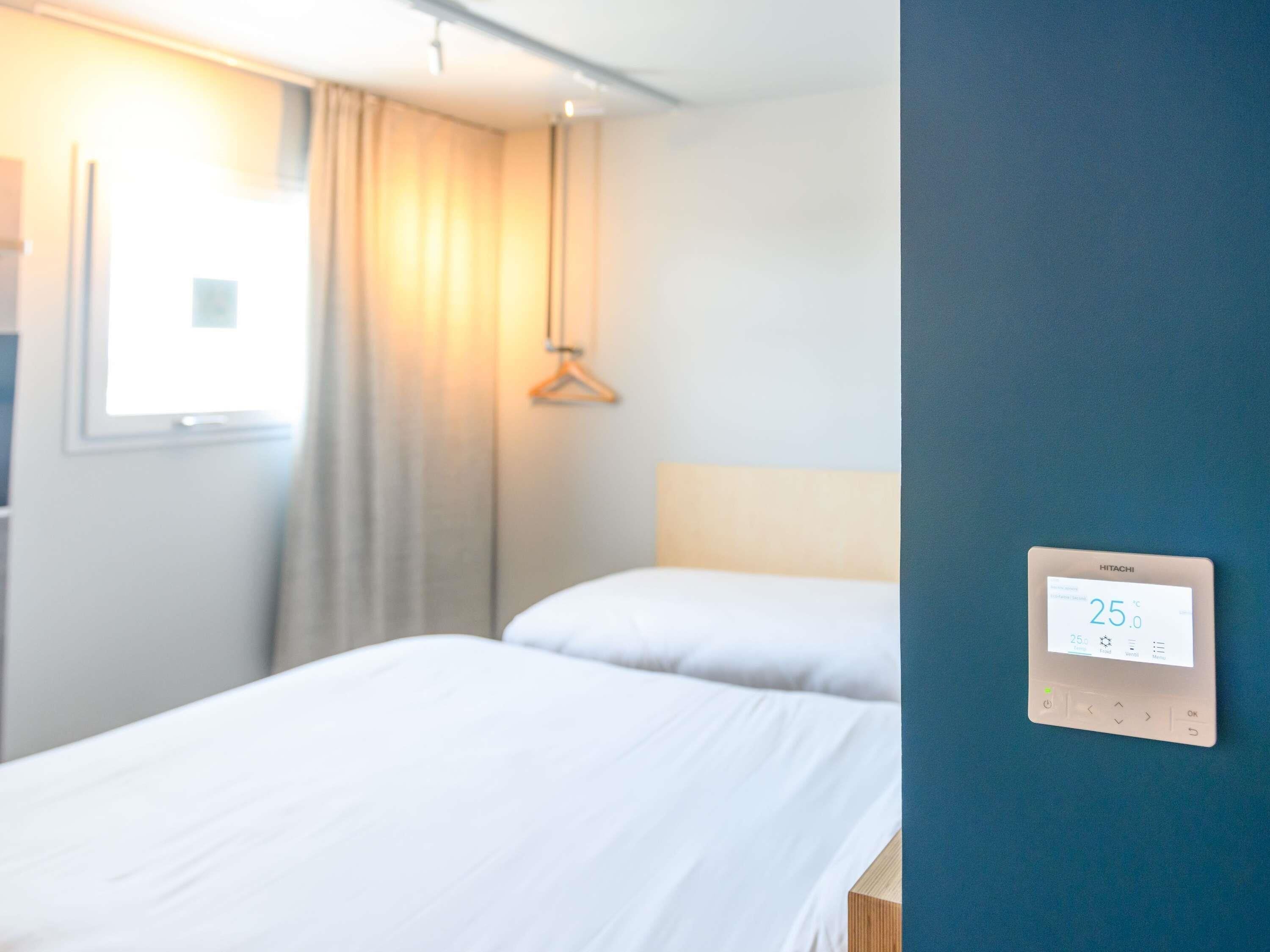 Habitación de hotel con dos camas, una ventana con cortinas, un termostato en la pared y una lámpara colgante.