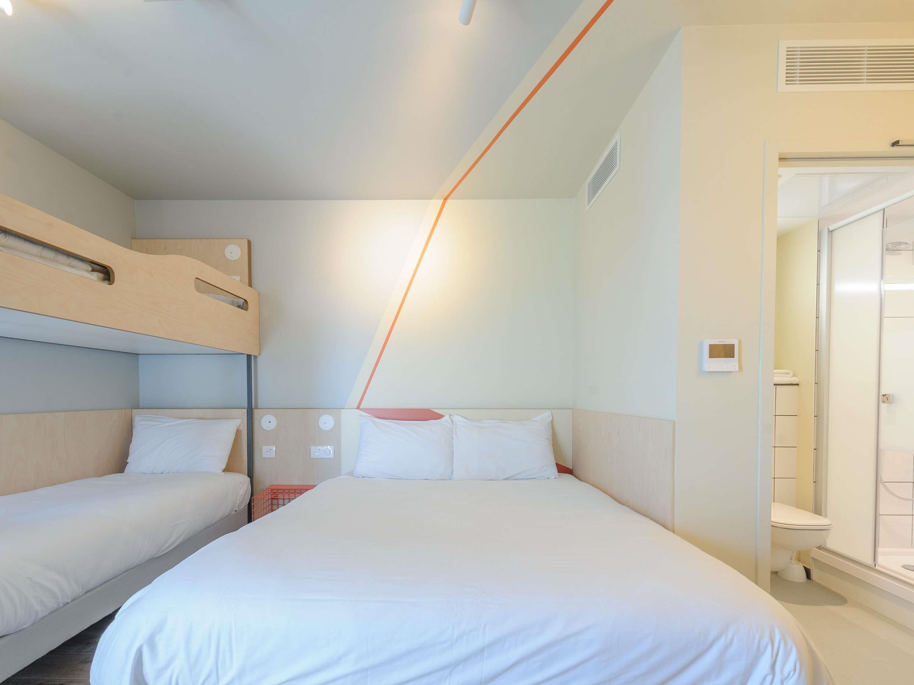 A hotel room with a bunk bed, a single bed, a shower area, and a thermostat on the wall.