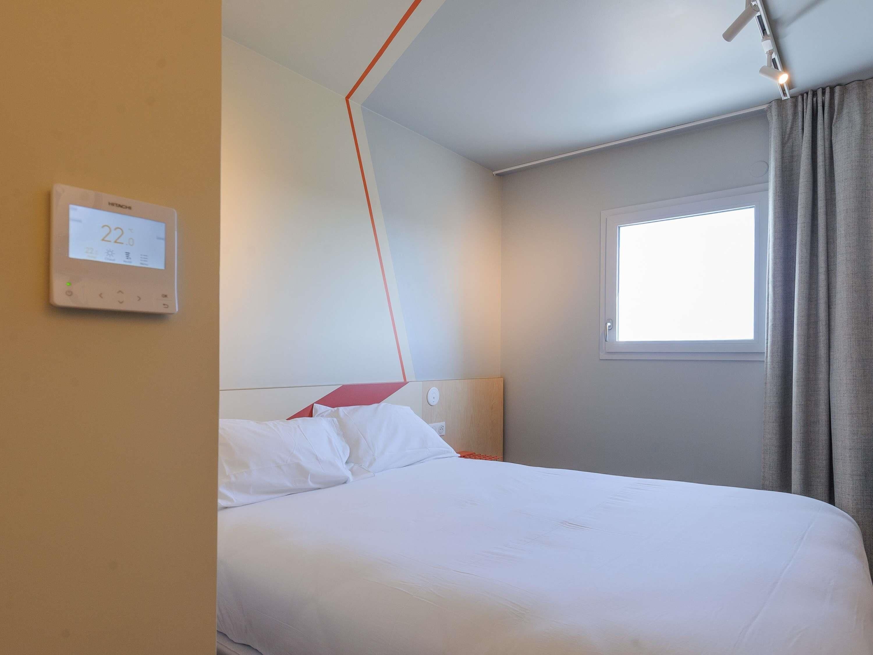 A hotel room with a bed, a thermostat, a window, and a curtain.