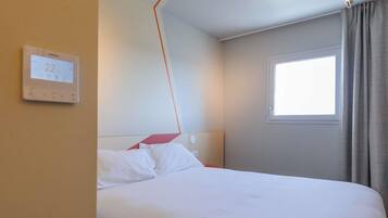 A hotel room with a bed, a thermostat, a window, and a curtain.