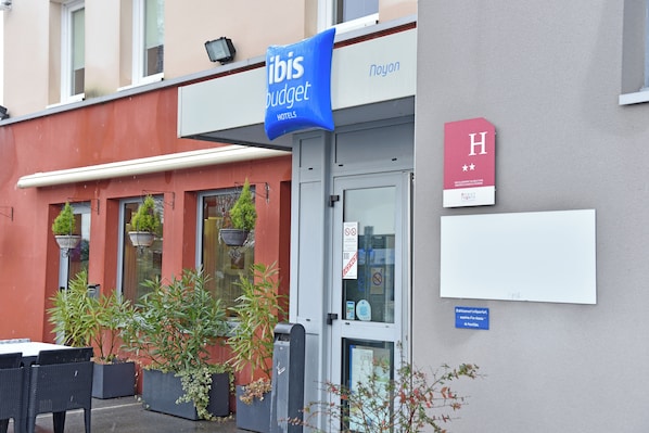 A hotel entrance with a blue banner reading "ibis budget hotels," a red sign with a white 'H' and stars, and a door with a glass panel.