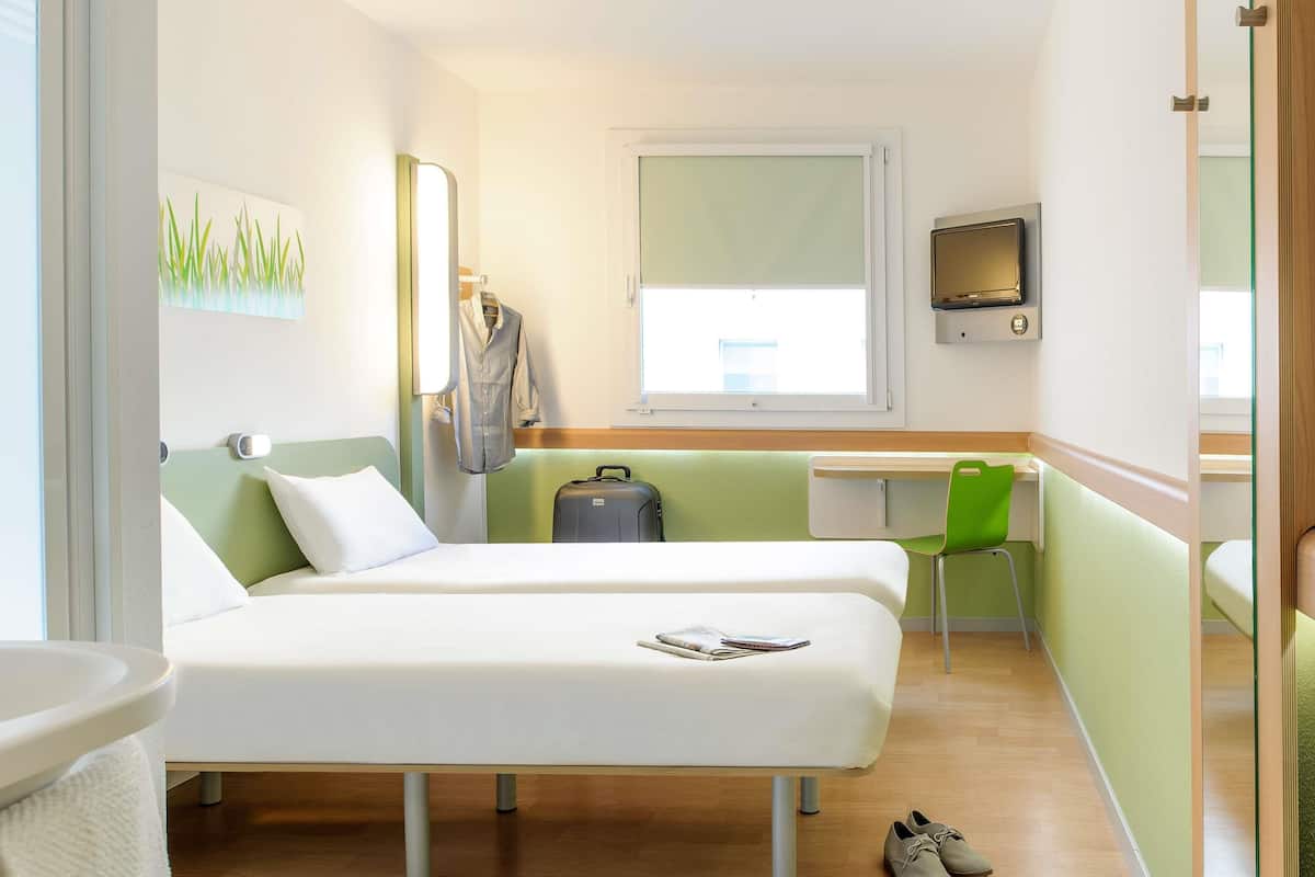 A hotel room with two beds, a desk, a chair, a suitcase, and a pair of shoes on the floor.