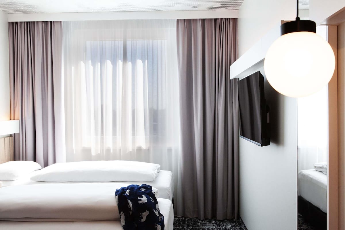 Habitación de hotel con cama, televisor montado en la pared y un amplio ventanal con cortinas.