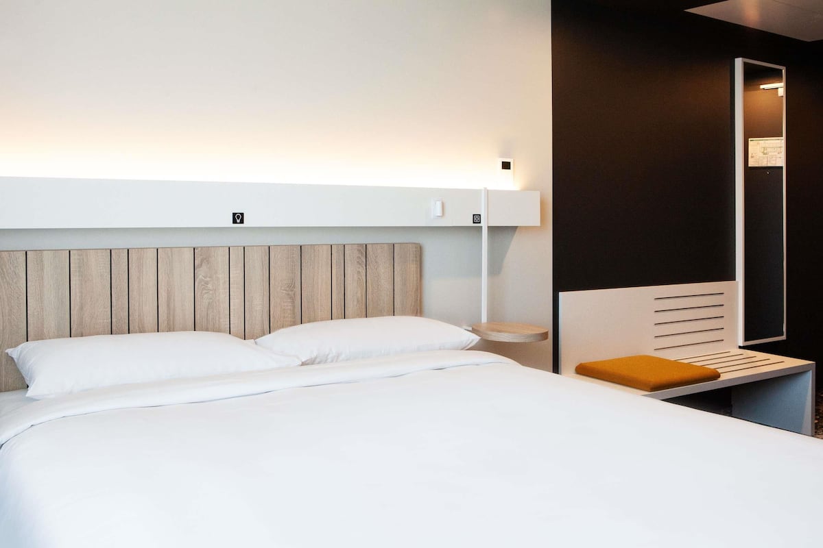 A modern hotel room with a wooden headboard, a white bed, a small wooden side table, and a large mirror.