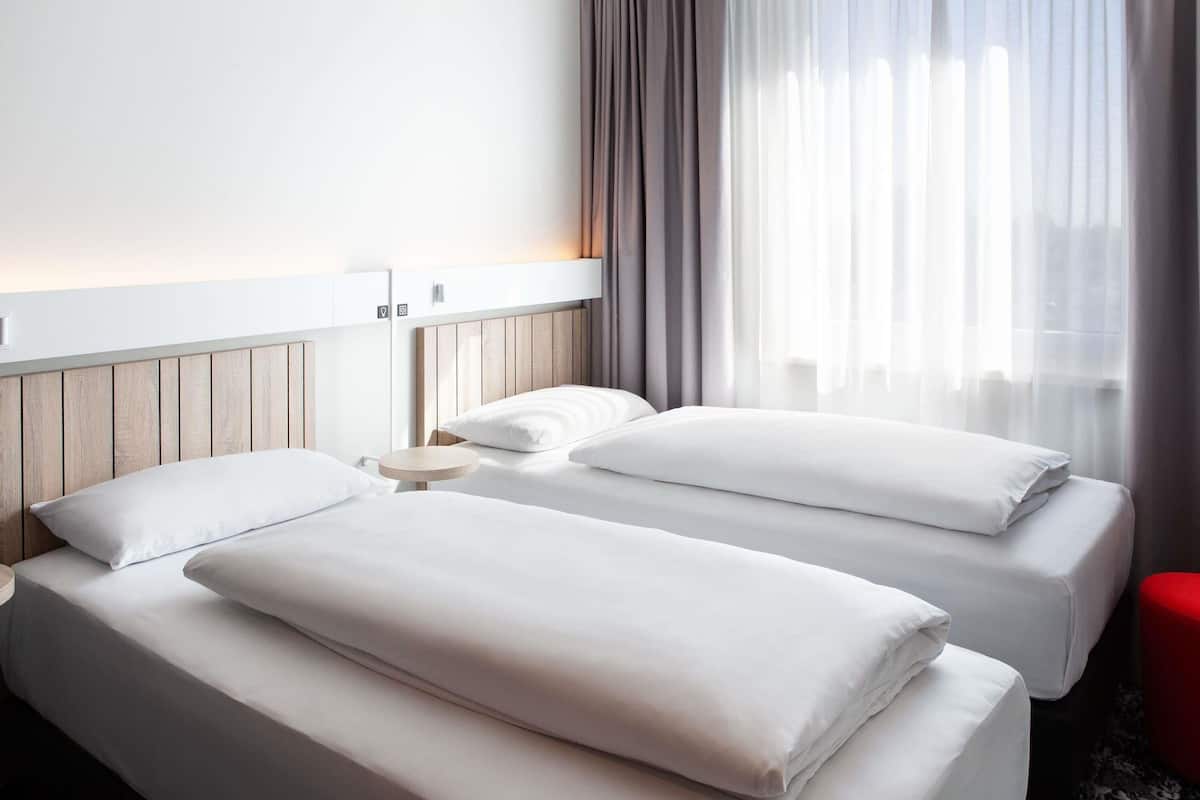 A hotel room with two beds, a round bedside table, a red stool, and a window with sheer curtains.