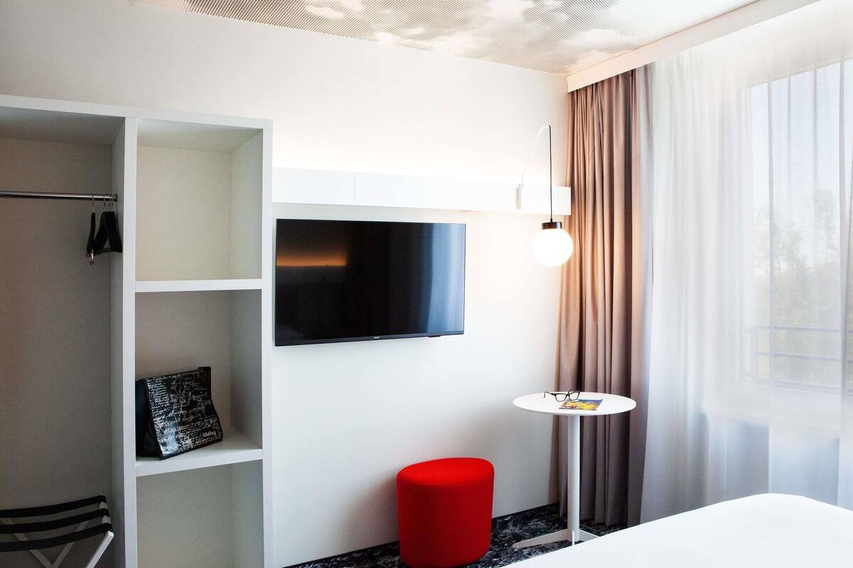 Habitación de hotel con cama, televisor montado en la pared, una mesita con vasos y una revista, un taburete rojo y un armario con estantes.