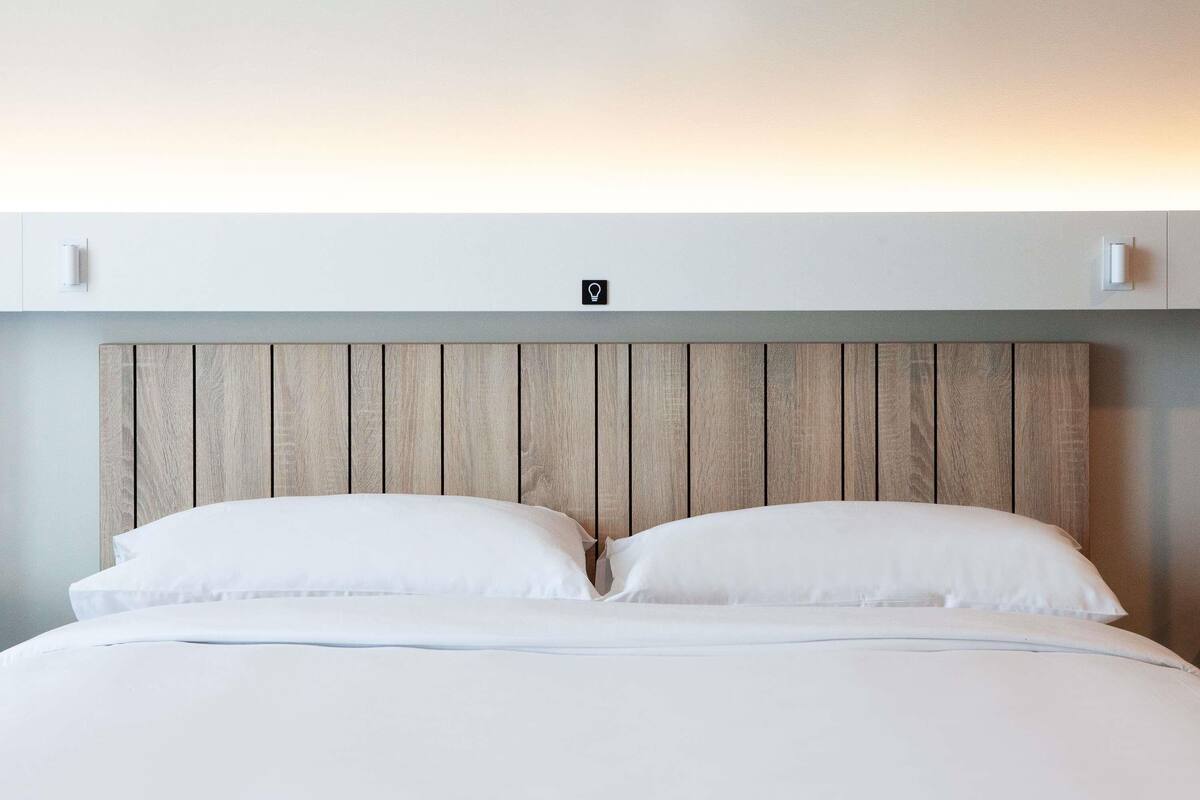 Una cama con cabecera de madera, ropa de cama blanca y un interruptor de luz en la pared.