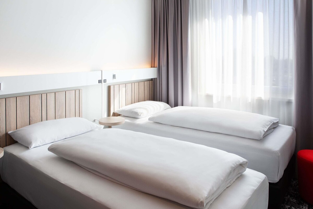 Habitación de hotel con dos camas, una mesita de noche redonda, un taburete rojo y una ventana con cortinas de gasa.