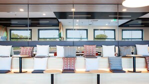Een moderne zithoek met meerdere tafels en banken, grote ramen en een plafond met lineaire lichtarmaturen.