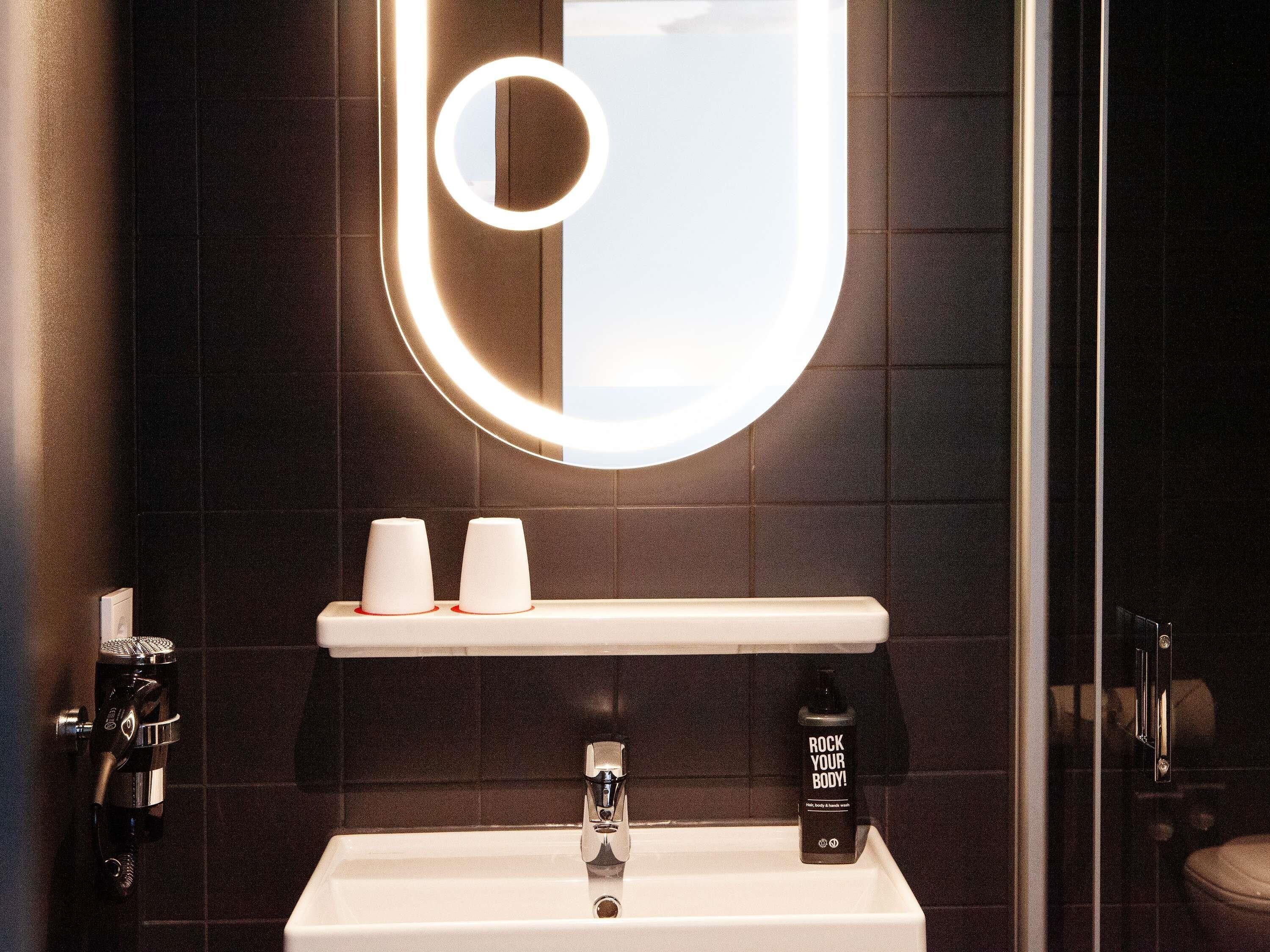 Un baño con un lavamanos blanco, un espejo con luz circular, dos tazas blancas en un estante y una botella de jabón de cuerpo.