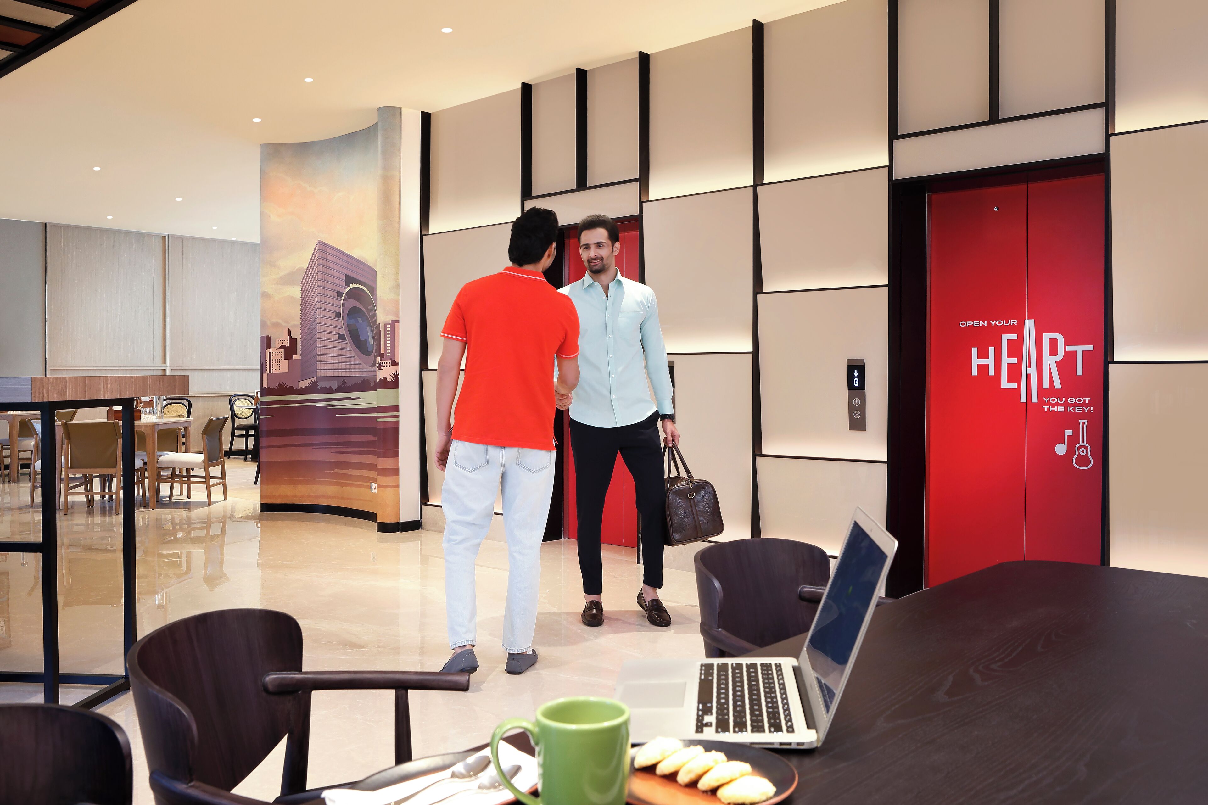 Un moderno vestíbulo de hotel con una pared roja que muestra el texto "OPEN YOUR HEART YOU GOT THE KEY". Se ven dos personas conversando, un mostrador de recepción y un área de descanso con sillas y una mesa.