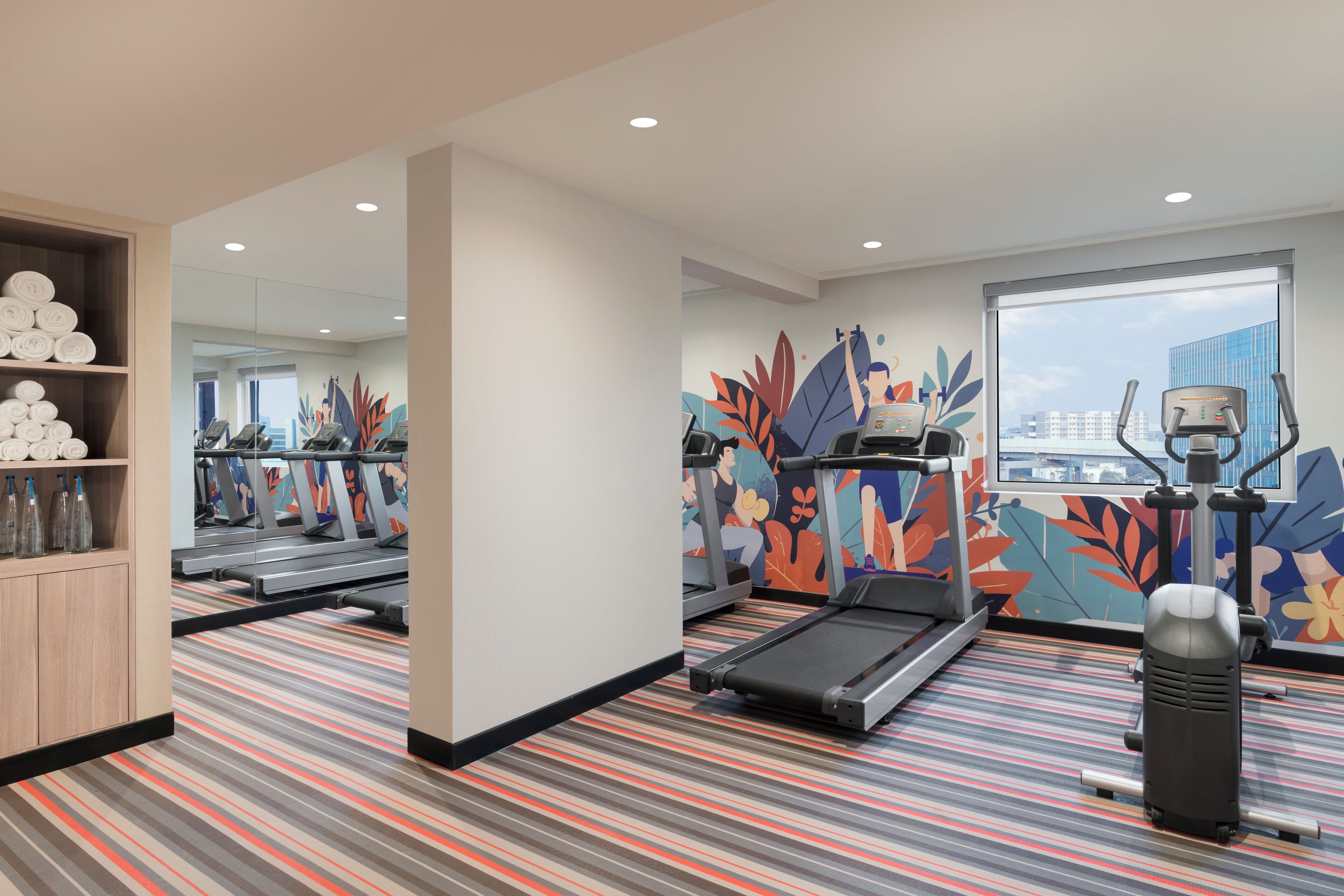 Une salle de sport équipée de tapis de course, d’un vélo d’appartement et d’un rameur. Des serviettes et des bouteilles d’eau sont disposées sur des étagères. La pièce est dotée d’un tapis à rayures et d’une grande fenêtre donnant sur la ville.