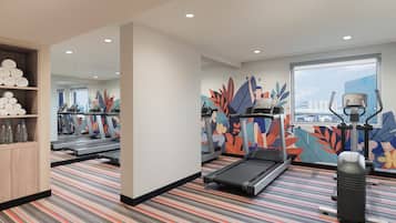 Une salle de sport équipée de tapis de course, d’un vélo d’appartement et d’un rameur. Des serviettes et des bouteilles d’eau sont disposées sur des étagères. La pièce est dotée d’un tapis à rayures et d’une grande fenêtre donnant sur la ville.